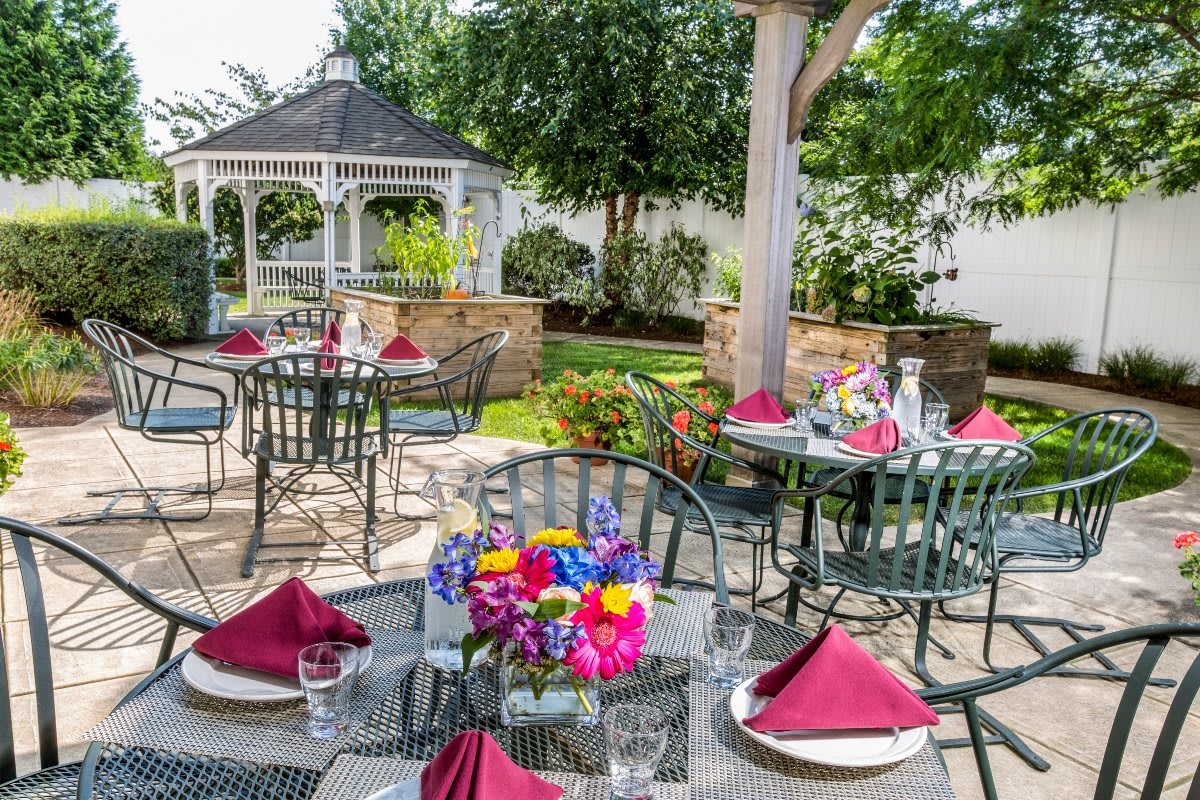 An elegant outdoor garden setting with tables adorned with flowers and burgundy napkins, surrounded by greenery and a gazebo.