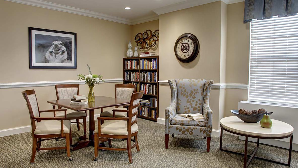 A cozy living room with a small table, chairs, bookshelf, and wall clock.
