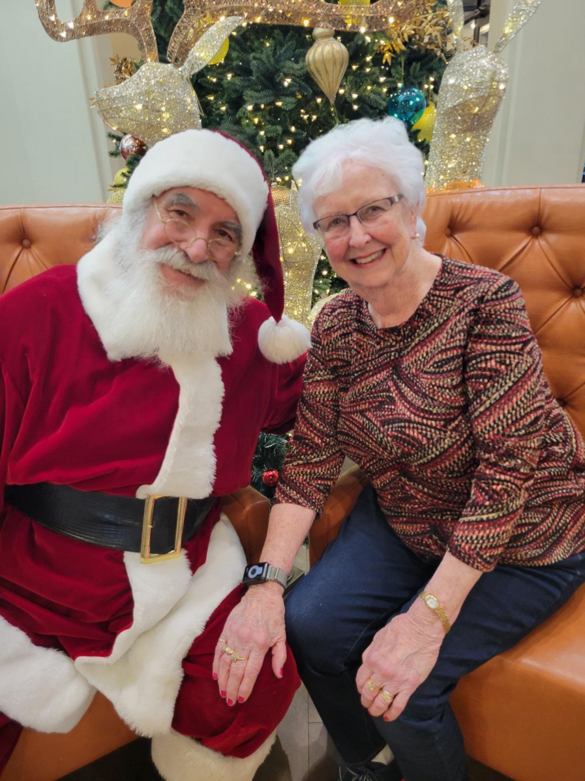 An elderly woman smiling while sitting next to Santa Claus in a festive setting with a decorated Christmas tree.