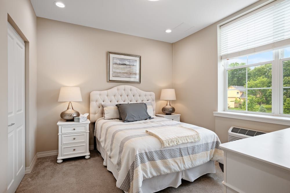 A cozy bedroom with neutral tones, featuring a bed, nightstands with lamps, and a window allowing natural light.