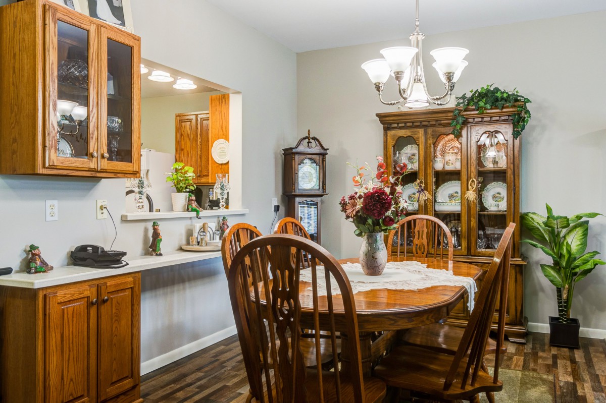 A cozy dining room with wooden furniture and a chandelier, featuring a table set for dining and a view into the kitchen.