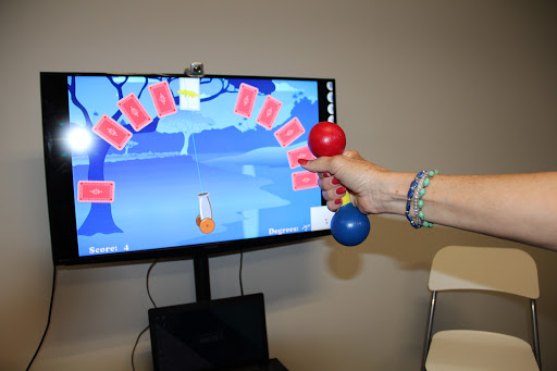 A person uses dumbbells in front of a screen displaying an interactive game related to physical fitness.