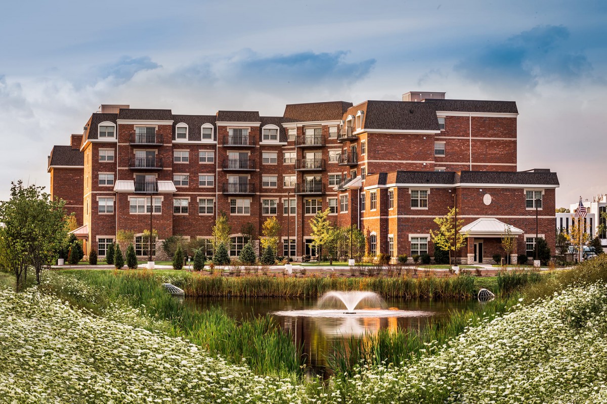 A residential building with a pond and fountain in front, surrounded by flowers and landscaping.