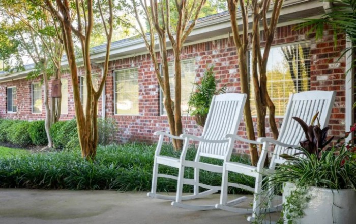 Two white rocking chairs on a patio with trees and a brick house in the background.
