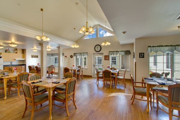 A bright dining room with wooden flooring, several tables and chairs, decorative lighting, and large windows.
