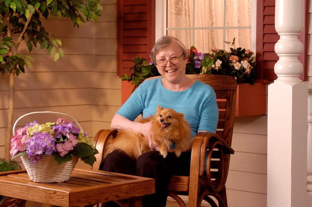 A woman smiles while sitting on a porch with a dog on her lap.