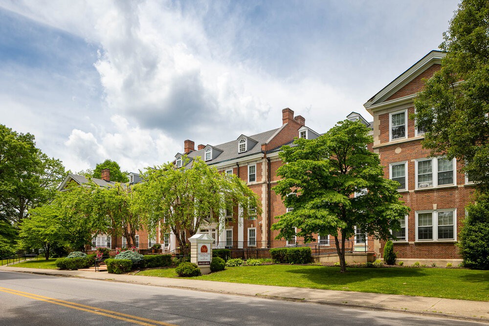 A picturesque brick building with a manicured lawn and trees lining the street under a cloudy sky.