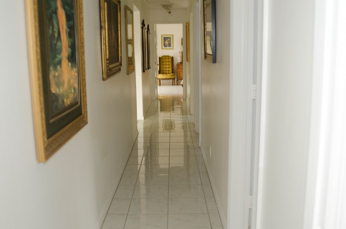 A long hallway with tiled flooring and artwork on the walls leading to a distant chair.