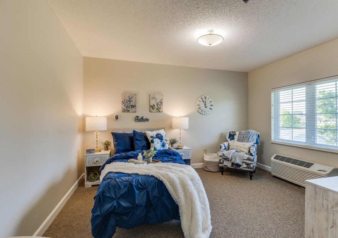 A well-decorated bedroom featuring a cozy bed with blue and white bedding, a comfortable chair, and warm lighting from lamps on either side.