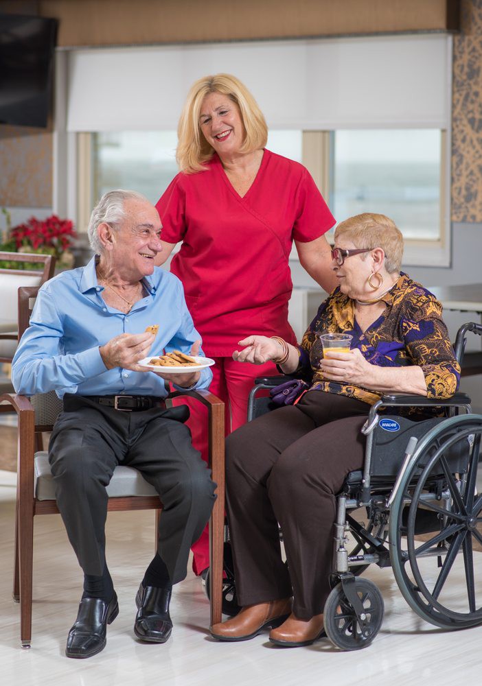 An elderly man and woman enjoying a meal with the support of a caregiver in a nursing home setting.