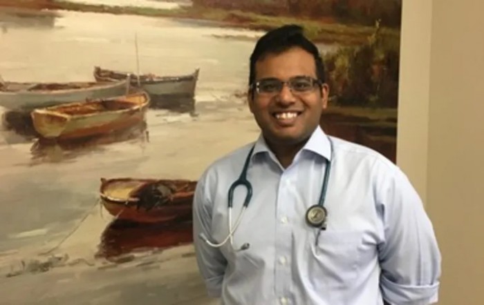 A smiling doctor stands in front of a painting featuring boats on the water.