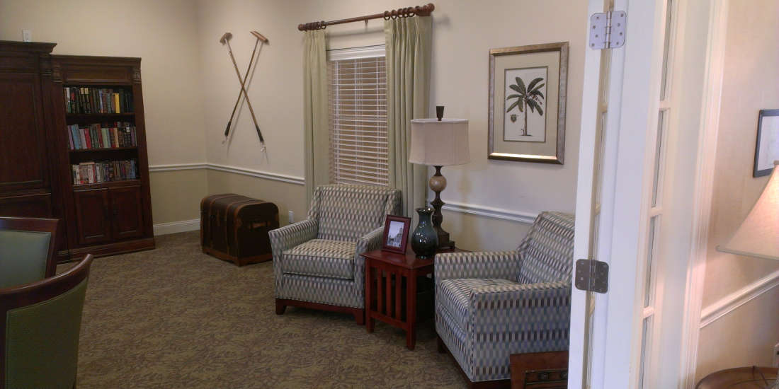 A cozy waiting room featuring two striped armchairs, a side table with a lamp, wall-mounted golf clubs, and palm tree artwork.