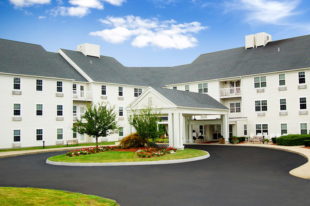 A large white residential-style building with a circular driveway and well-maintained landscaping under a partly cloudy sky.