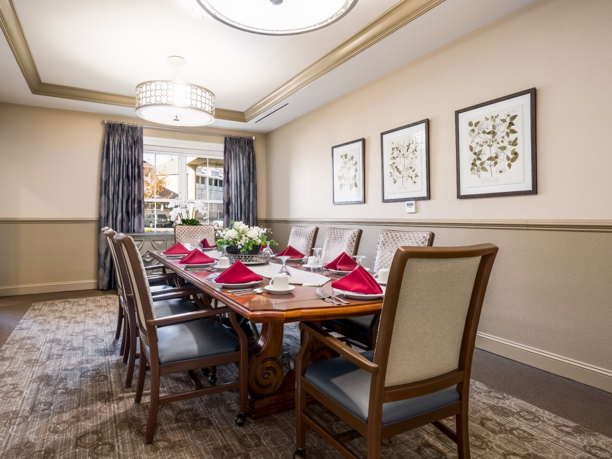 A formal dining room with an elegantly set table, featuring red napkins and floral centerpieces, surrounded by comfortable chairs and decorated with framed artwork.