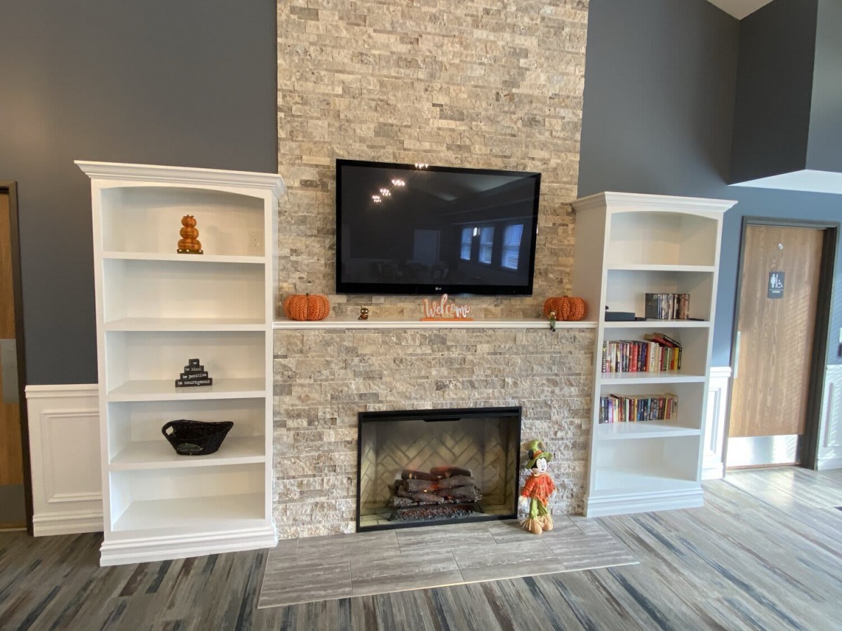 A cozy living room setup with a central stone fireplace, a mounted TV, and built-in shelves decorated with books and pumpkins.