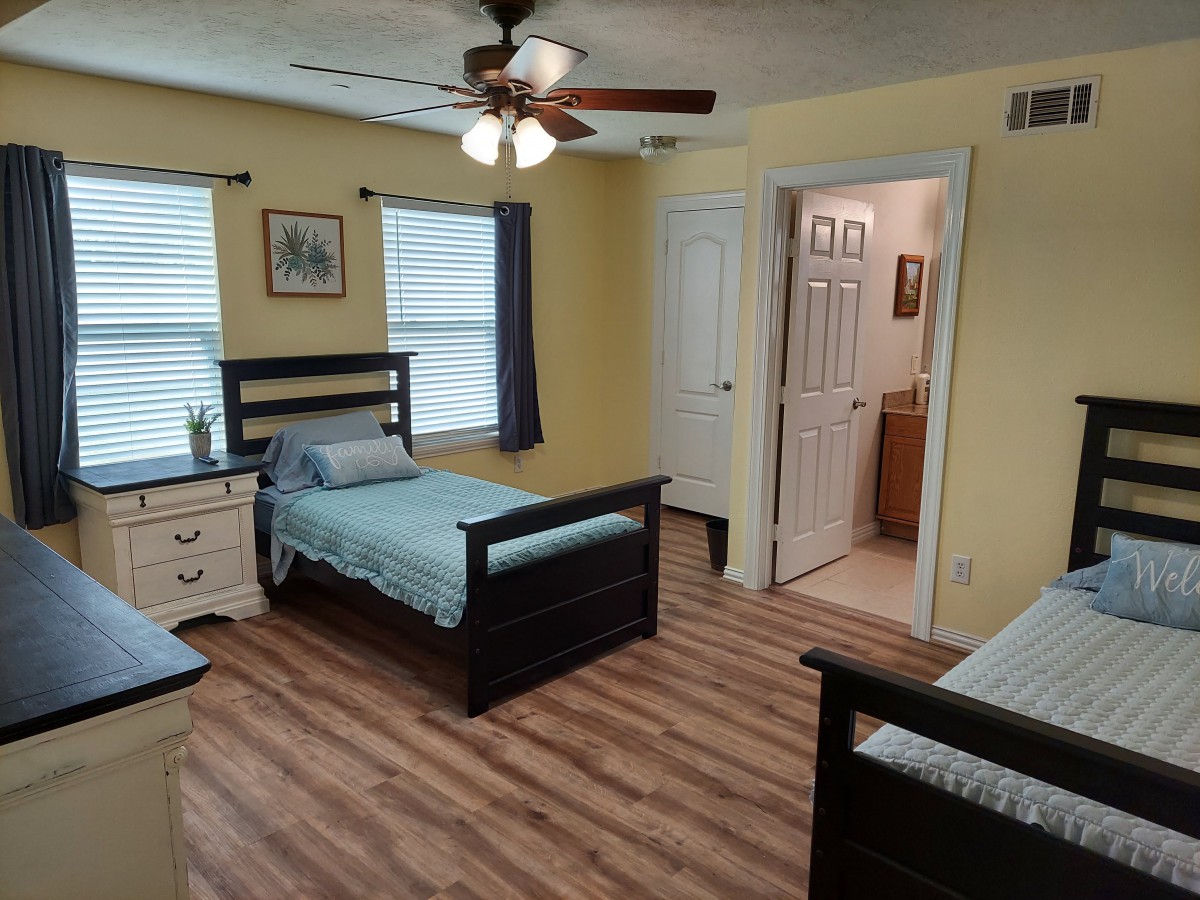 A cozy bedroom featuring twin beds, wooden flooring, and a ceiling fan. The room has yellow walls, two windows with curtains, a dresser, and an adjoining bathroom.