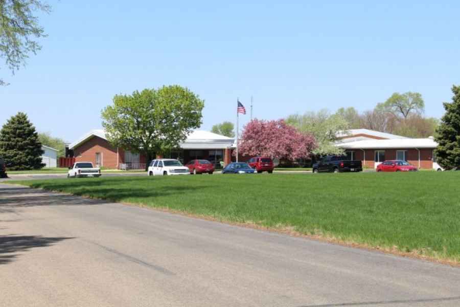 A building with a flag and cars parked in front, surrounded by trees and grass on a sunny day.