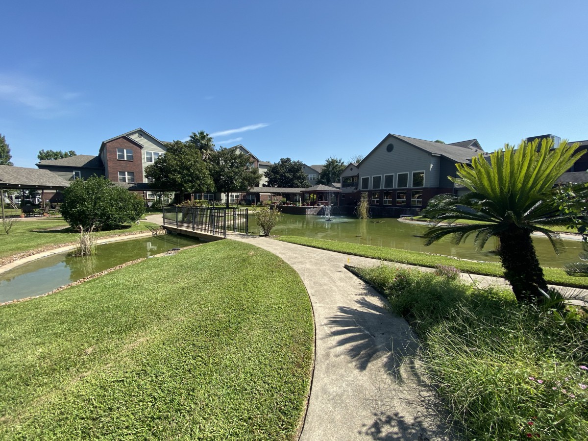 A scenic view of a landscaped garden featuring a winding pathway, pond, bridge, and several buildings under a clear blue sky.