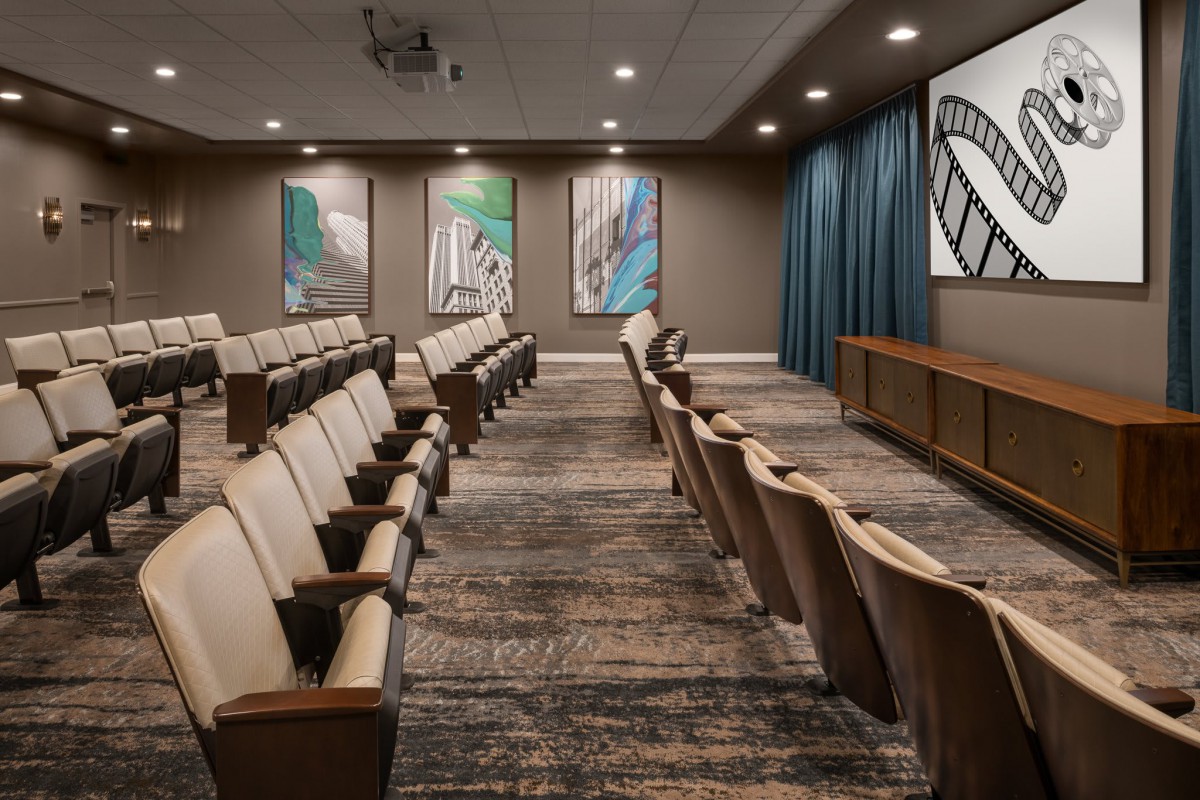 An empty auditorium with rows of seating facing a projector screen, adorned with abstract artwork on the walls.