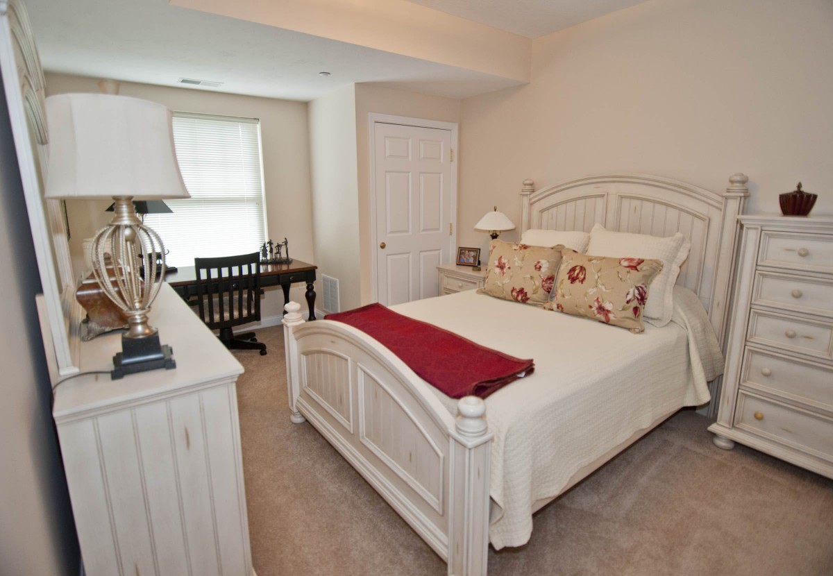 A cozy bedroom with a neatly made bed, decorated with pillows and a red throw, featuring a desk and chair in the corner.