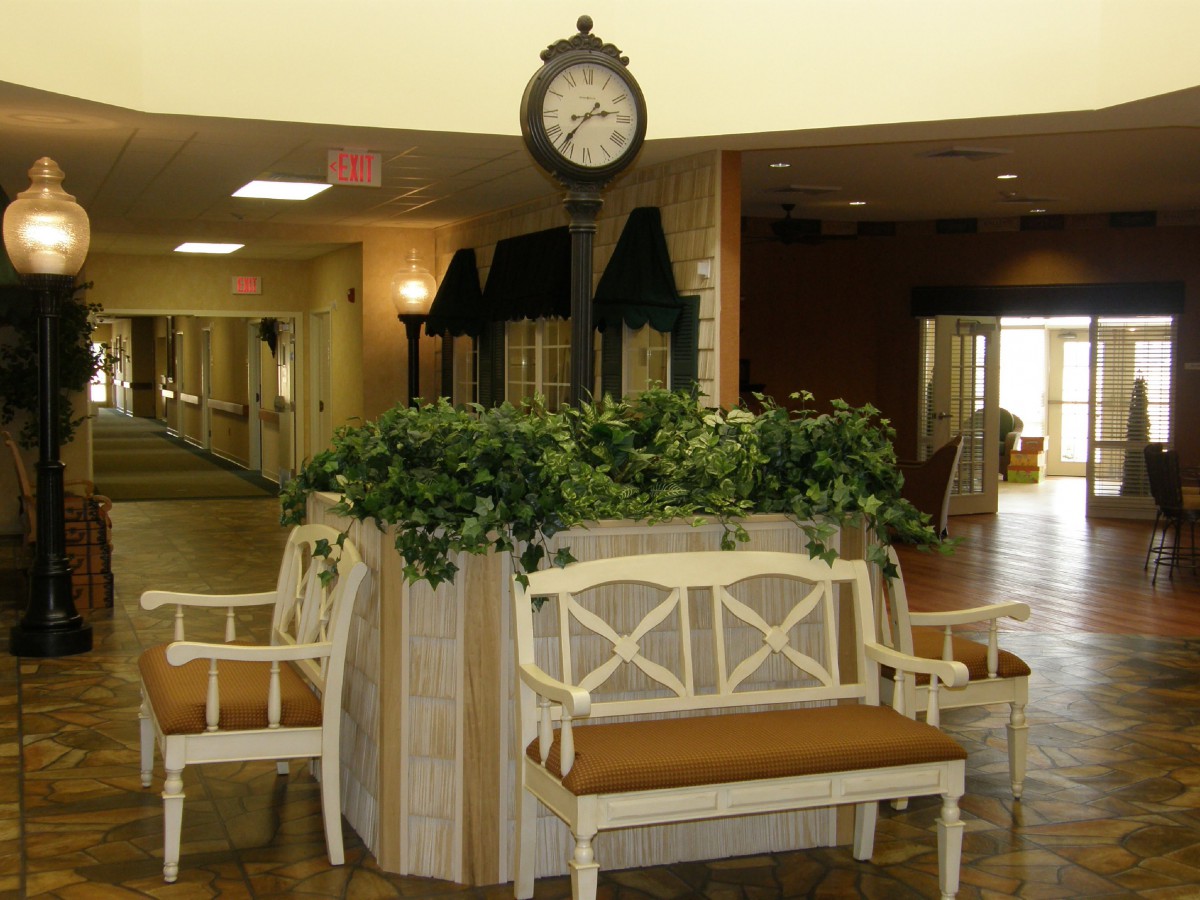 A lobby interior featuring a central seating area with benches and a decorative clock above lush plants.