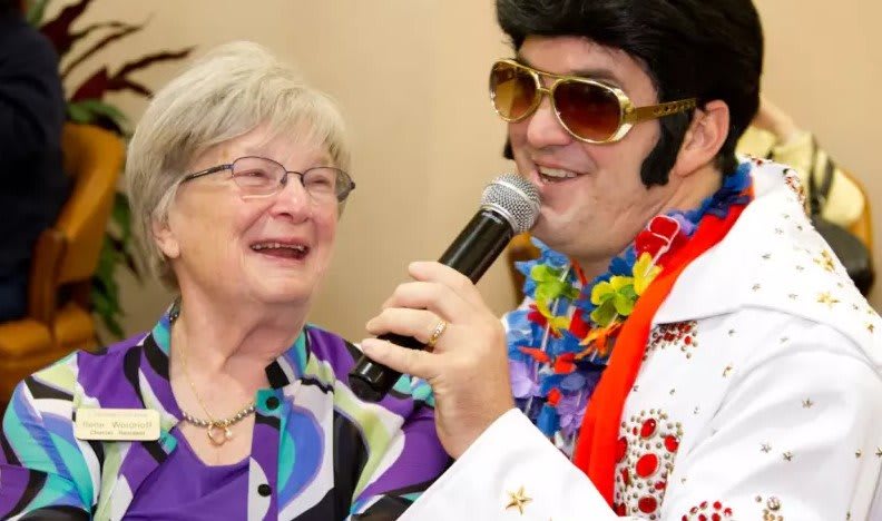 An elderly woman shares a joyful moment with a man dressed in a vibrant costume, holding a microphone.