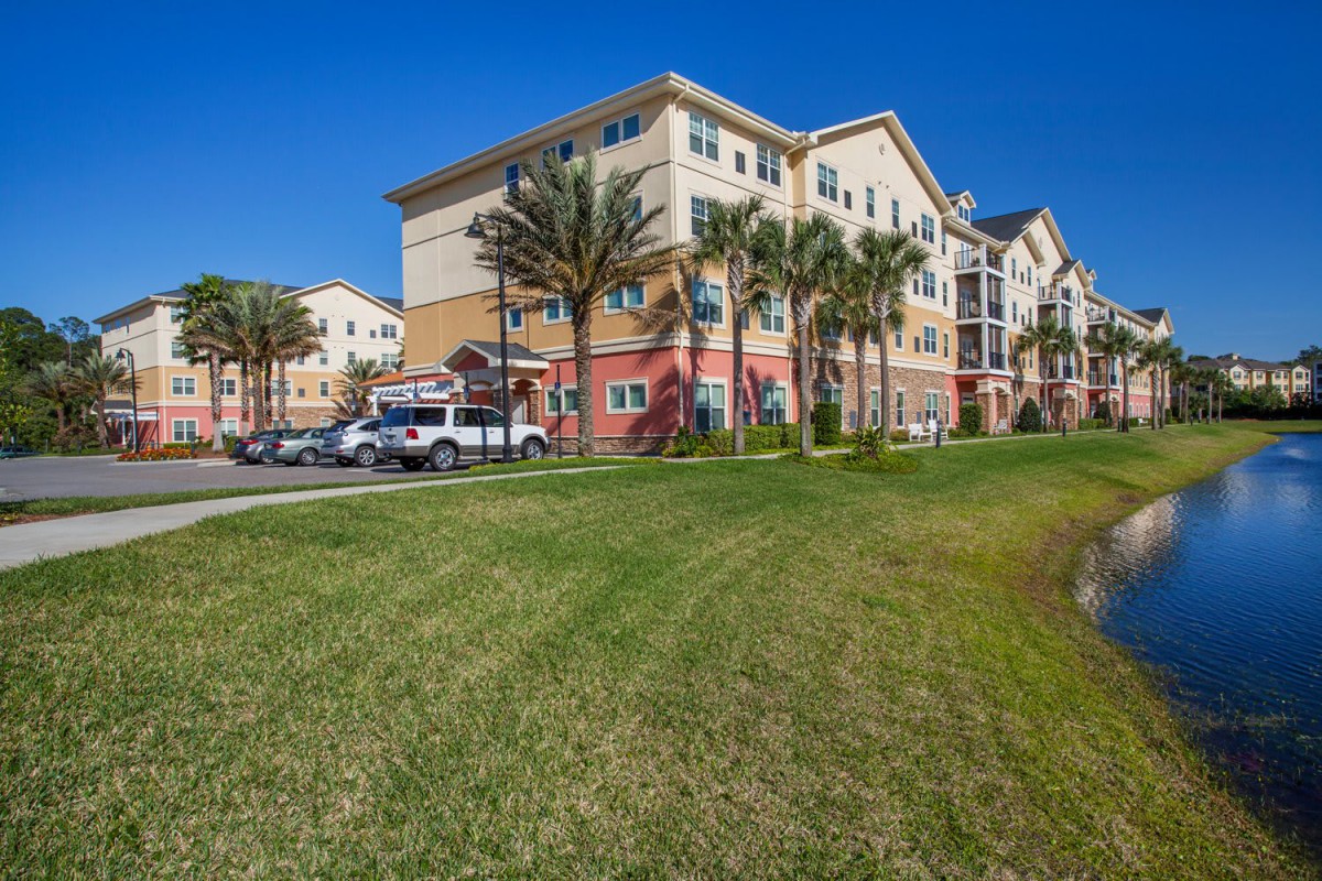 A multi-story apartment building with palm trees and a parking lot on a sunny day.