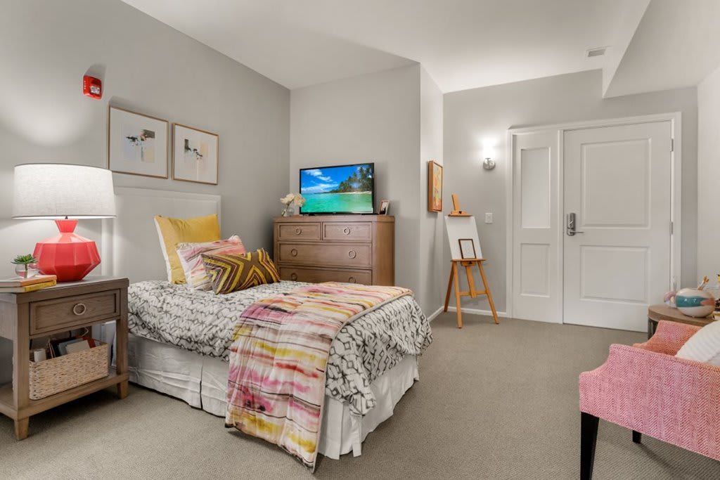 A cozy bedroom featuring a single bed with colorful bedding, a television on a dresser, and wall art.