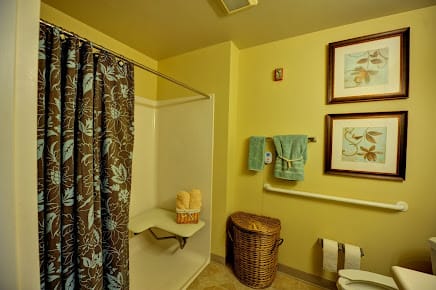 A cozy bathroom with yellow walls, featuring a shower with a patterned curtain, neatly hung towels, a wicker basket, and framed artwork on the wall.
