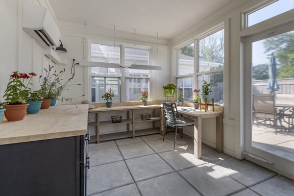 A bright sunroom filled with natural light, featuring large windows and a minimalist workspace adorned with plants.
