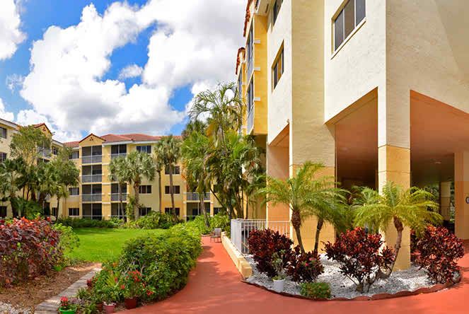 A view of an apartment complex with a landscaped pathway and palm trees under a partly cloudy sky.