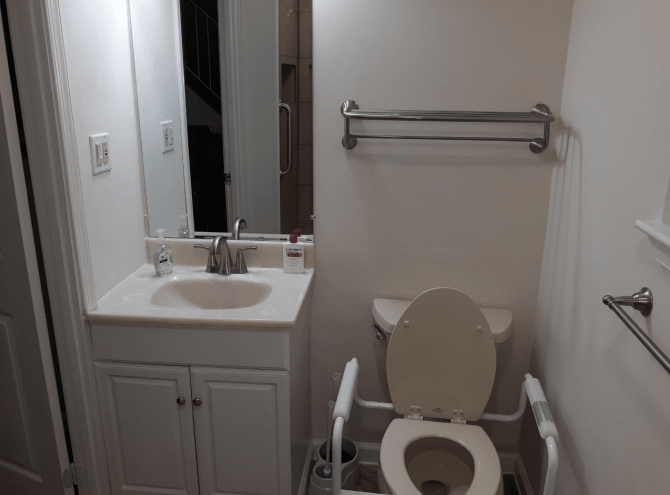 A bathroom with a vanity sink, mirror, and an accessible toilet equipped with handrails.