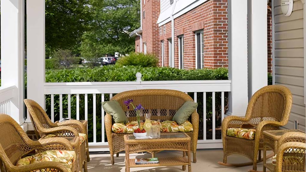 A cozy patio with wicker furniture and colorful cushions, set against a backdrop of a red brick building and lush greenery.
