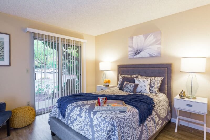 A cozy bedroom with a neatly made bed, featuring decorative pillows and a throw blanket, illuminated by a bedside lamp and natural light from large sliding glass doors.