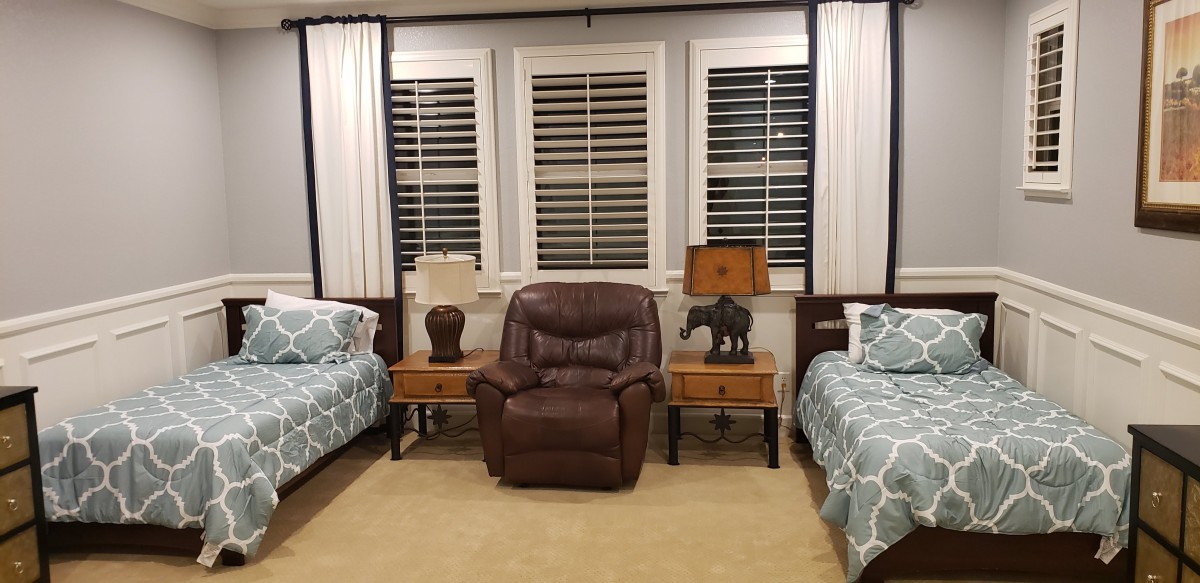 A cozy bedroom with twin beds and a recliner in between, flanked by nightstands and lamps. The windows are adorned with shutters and curtains.