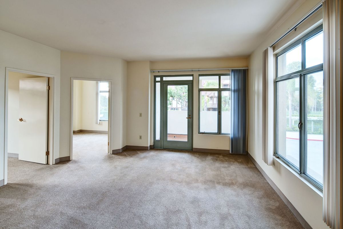 An empty living room with large windows and a glass door, featuring carpet flooring.