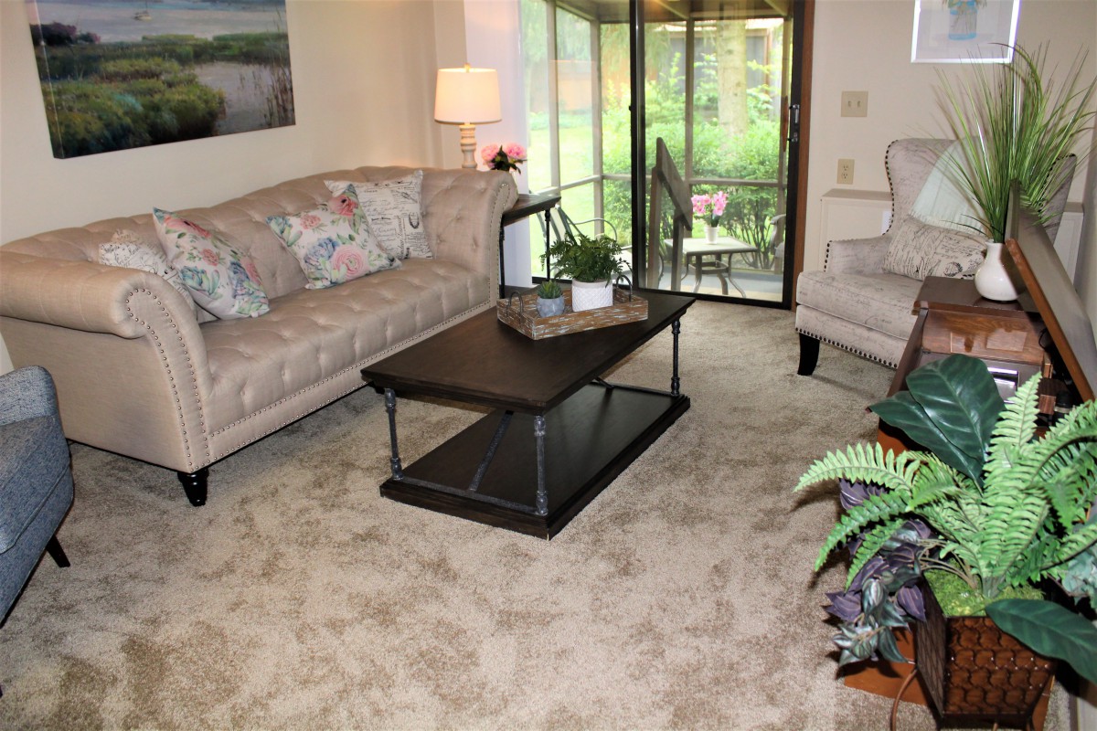 A cozy living room with a floral sofa, central coffee table, and a sliding glass door leading to an outdoor view.