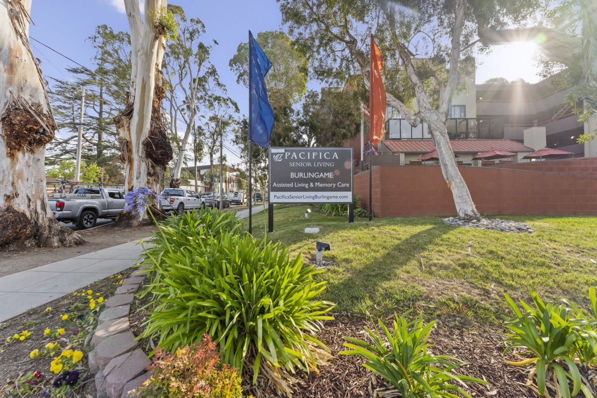 An outdoor view of an apartment complex sign surrounded by trees and landscaping, with flags and parked cars nearby.