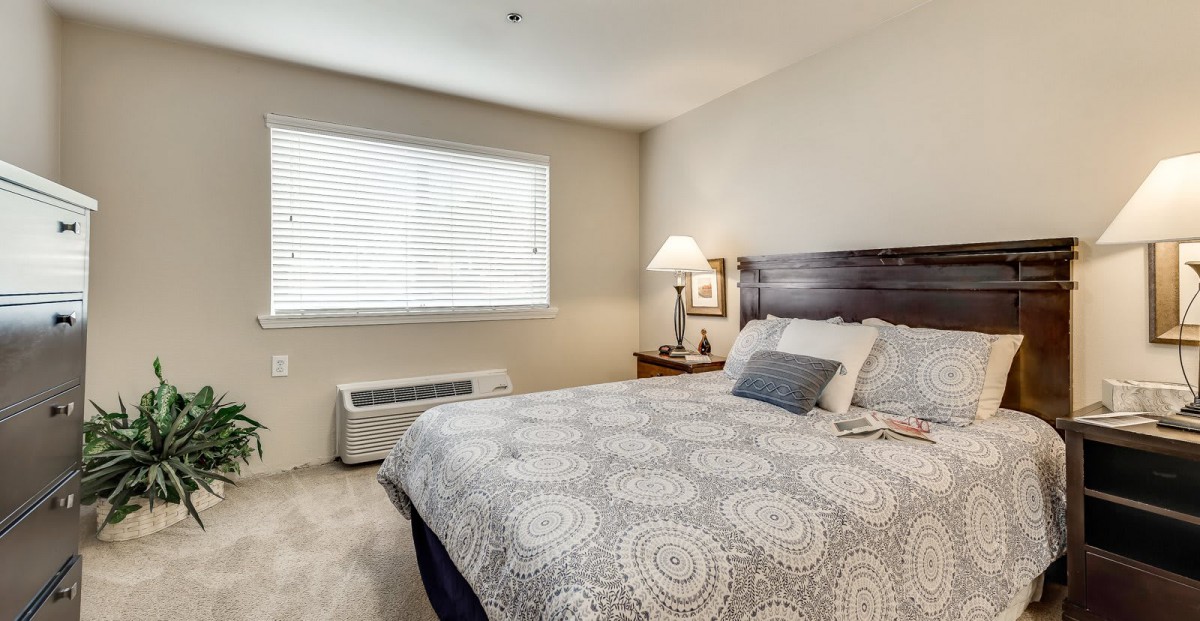 A cozy bedroom with a neatly made bed, bedside lamp, and a window providing natural light.