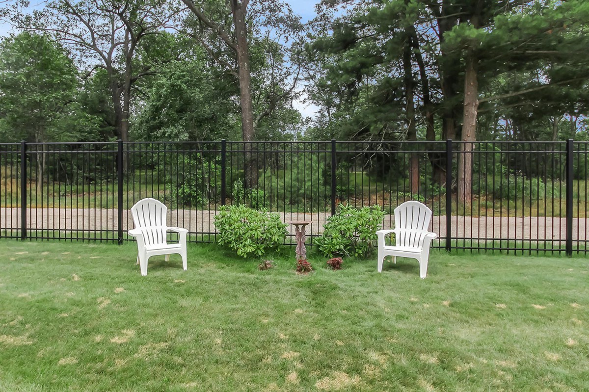 Two white chairs in a garden setting alongside a black fence and lush greenery.