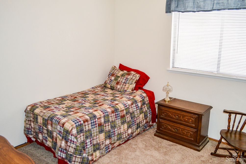 A small bedroom with a single bed featuring a colorful quilt, a wooden nightstand with a lamp, and a window with blinds.