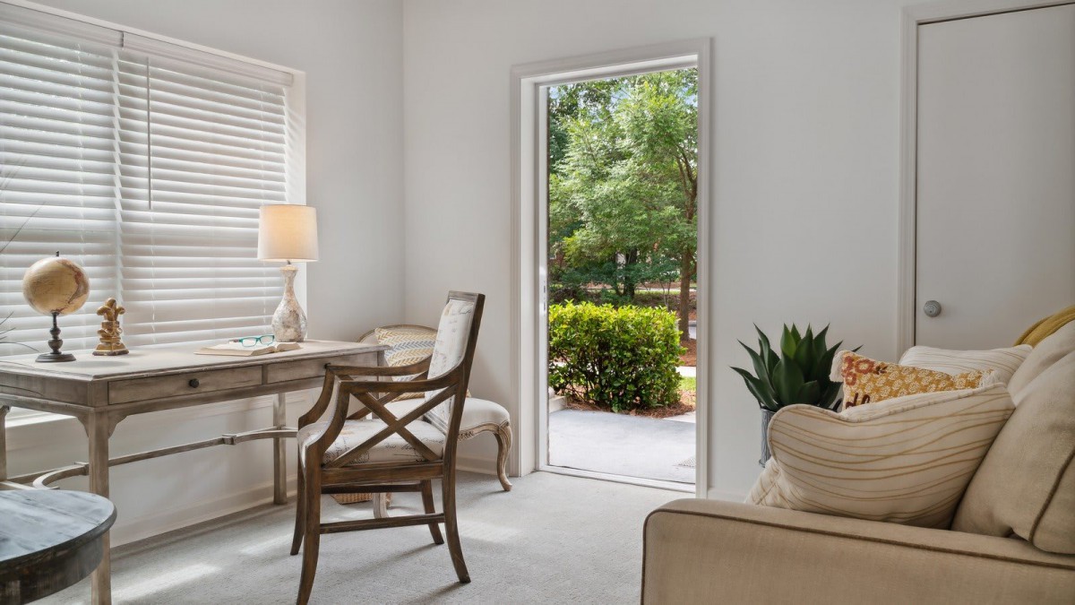 A cozy home office with a desk and chair, featuring a lamp and an indoor plant, overlooking a garden through an open door.