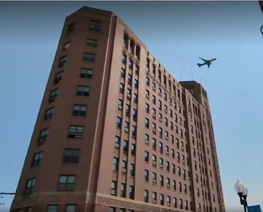 An airplane flies above a tall brick building against a clear blue sky.