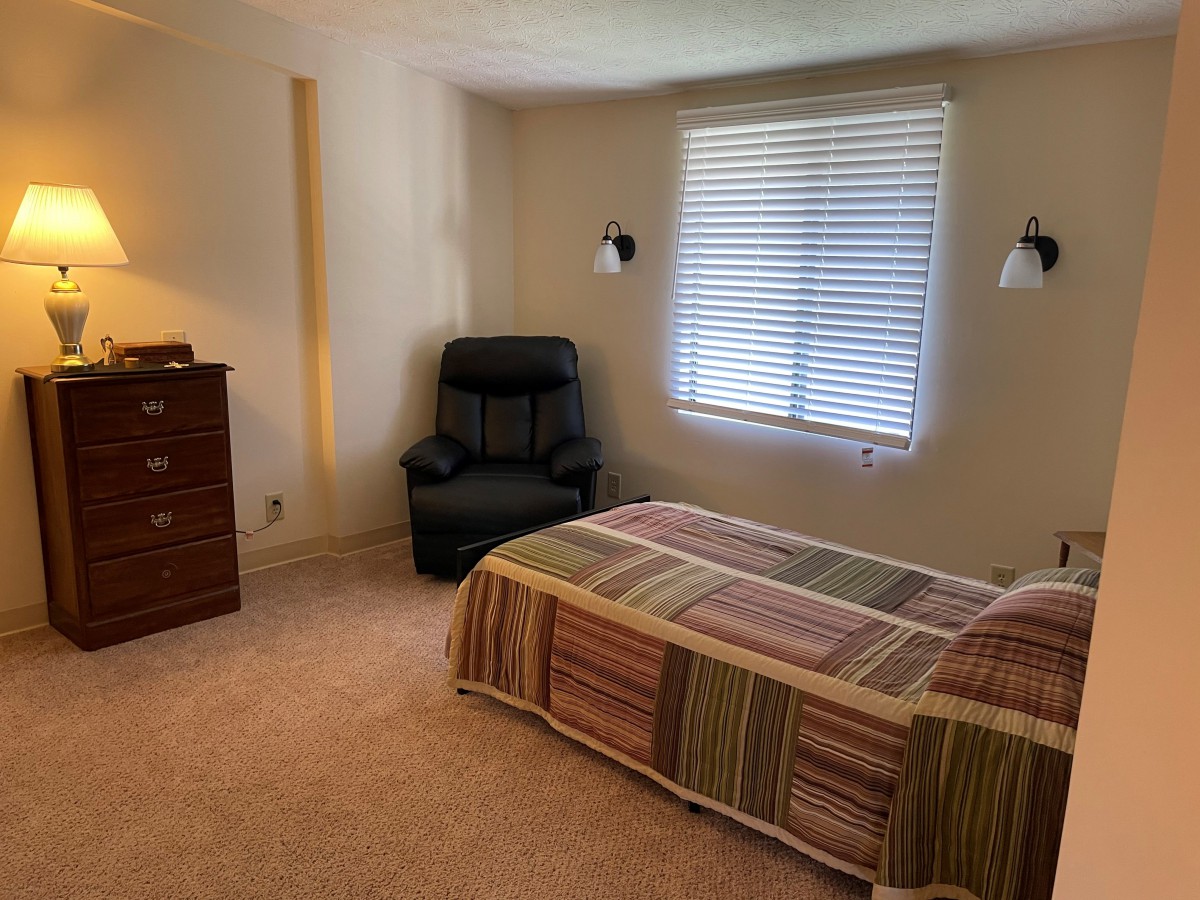 A cozy bedroom with a single bed, a recliner chair, a dresser, and natural light coming through the window.