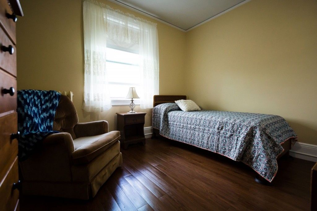 A simple bedroom with a single bed, an armchair, and a window letting in natural light.