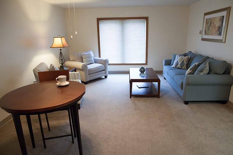 A cozy living room with a sofa, armchair, coffee table, and lamp, featuring a large window and carpeted floor.
