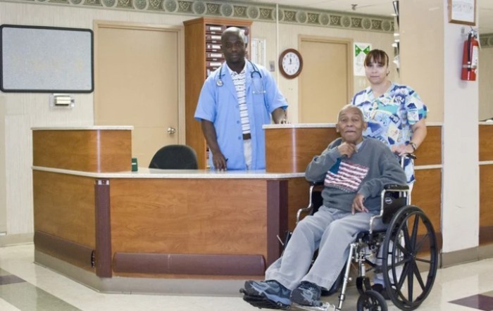 A doctor and nurse stand behind a patient in a wheelchair at a hospital reception area.
