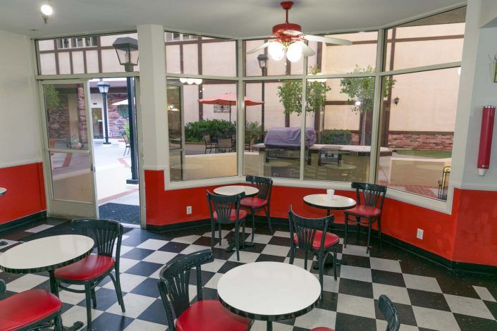 A cozy cafe with checkered flooring and red accents features several empty tables and chairs near large windows.
