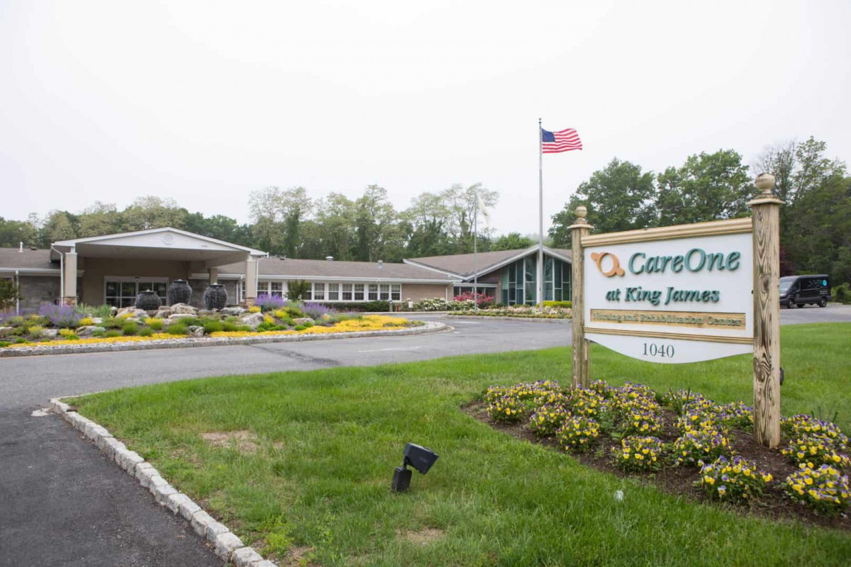 The image shows a healthcare facility with a prominent sign and an American flag in the front, surrounded by greenery.