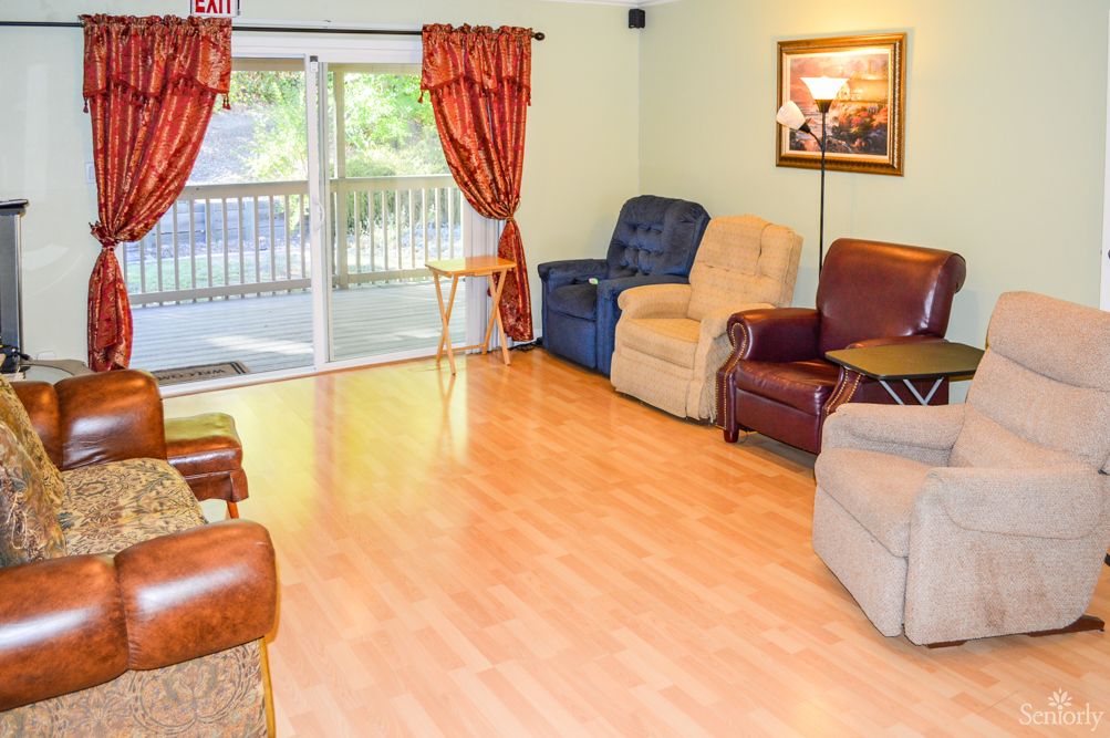 A cozy living room with hardwood flooring, featuring several recliner chairs and a large sliding glass door with red curtains letting in natural light.